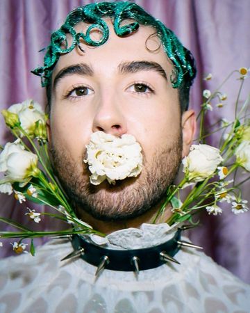 Adult male with glittery green hair and white flower in mouth poses with flower collar against purple backdrop in studio portrait.