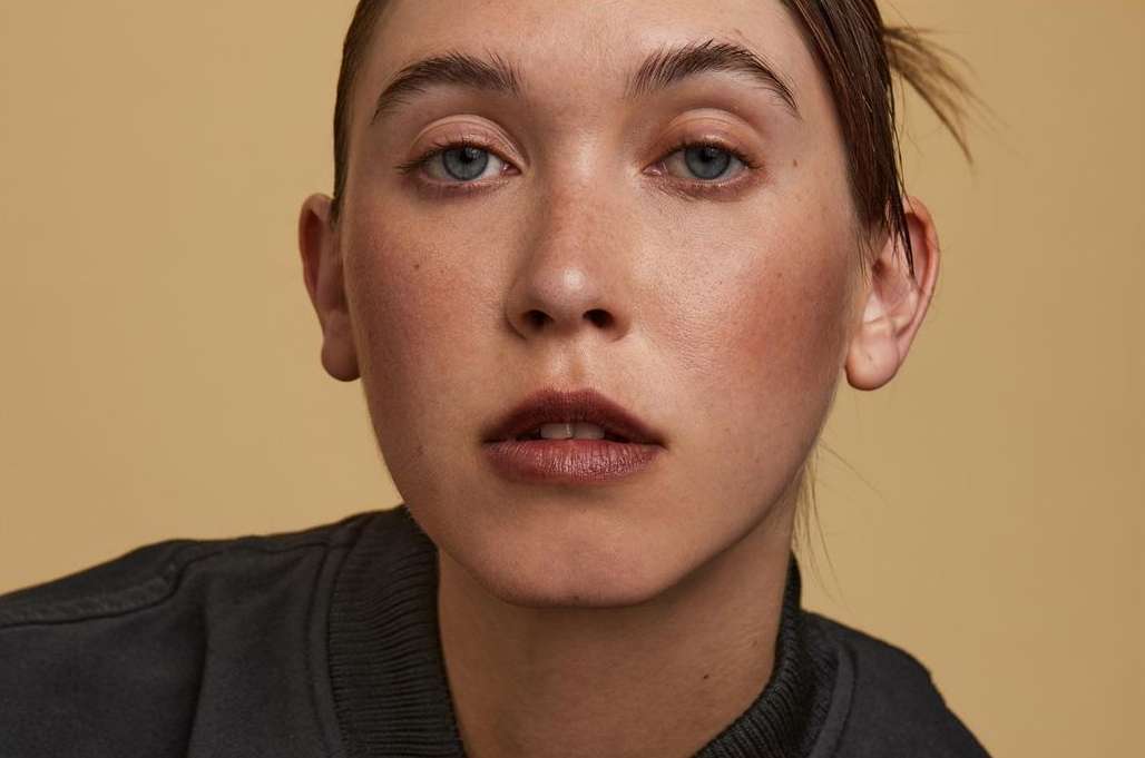 Close-up portrait of a young adult woman with light skin, wearing a dark top, against a plain tan backdrop, with soft, diffused lighting.