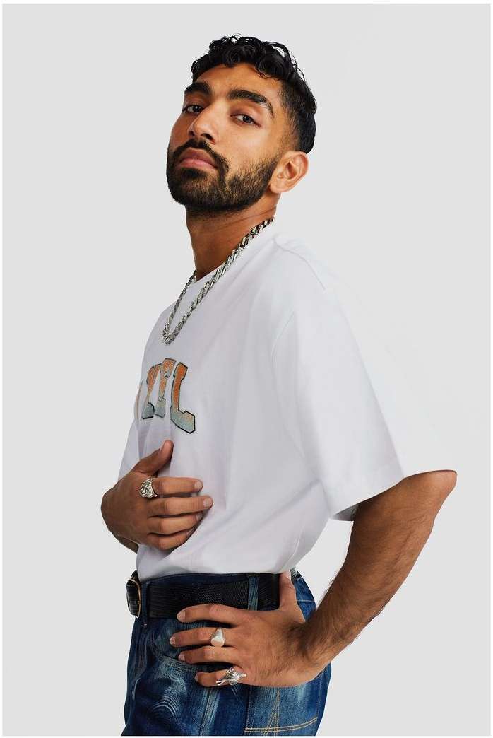 Portrait of an adult male with a beard wearing a white t-shirt, jeans, and jewelry in a studio setting with a neutral background.