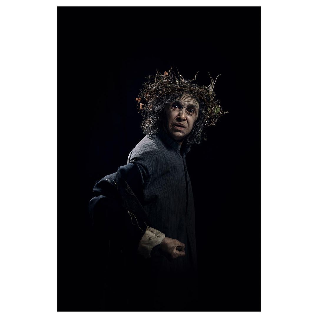 Dramatic portrait of an adult male with long hair and a crown of twigs, posing in a dark setting with theatrical lighting.