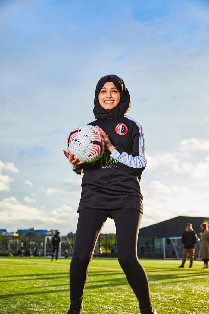 Female soccer player in hijab holding a soccer ball on a green sports field on a sunny day with people in the background.