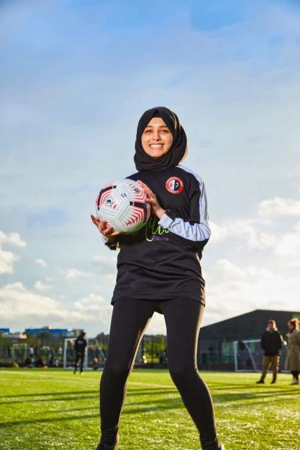 Female soccer player in hijab holding a soccer ball on a green sports field on a sunny day with people in the background.