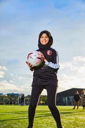 Female soccer player in hijab holding a soccer ball on a green sports field on a sunny day with people in the background.