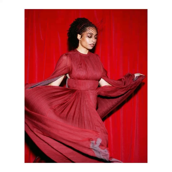 Woman in flowing burgundy gown with veil poses against vibrant red draped backdrop in fashion portrait. Elegant, formal attire, evening wear.