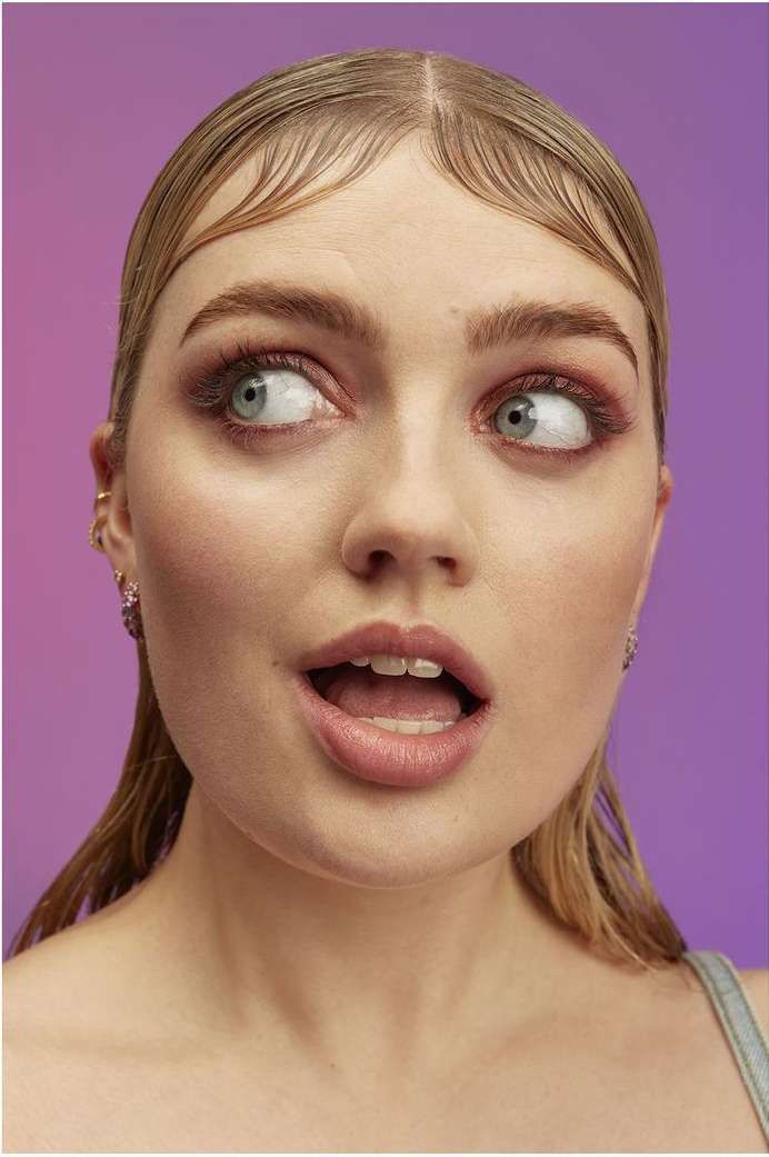 Close-up portrait of a young adult female with light blonde hair, light skin and pink eye shadow against a blended purple background.
