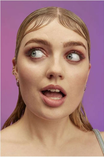 Close-up portrait of a young adult female with light blonde hair, light skin and pink eye shadow against a blended purple background.
