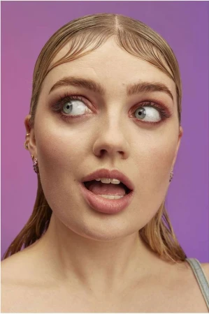 Close-up portrait of a young adult female with light blonde hair, light skin and pink eye shadow against a blended purple background.
