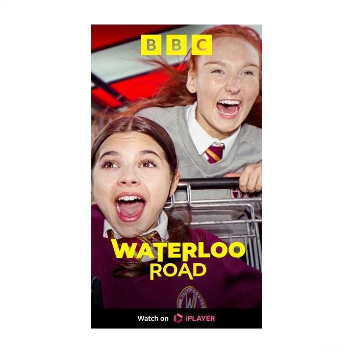 Two smiling students wearing school uniforms are riding in a shopping cart against a red background, promoting BBC's 'Waterloo Road'.