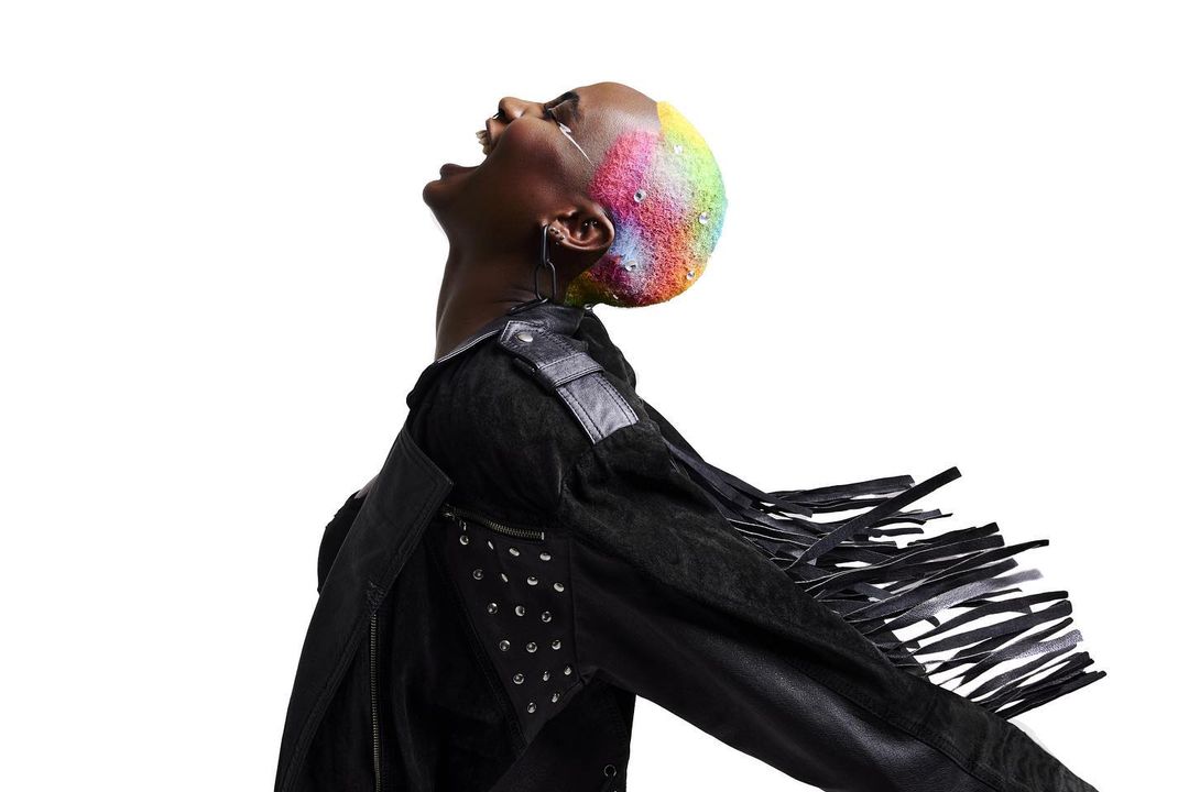 Side profile of a black adult female with rainbow dyed short hair wearing a fringed black leather jacket against a stark white background.