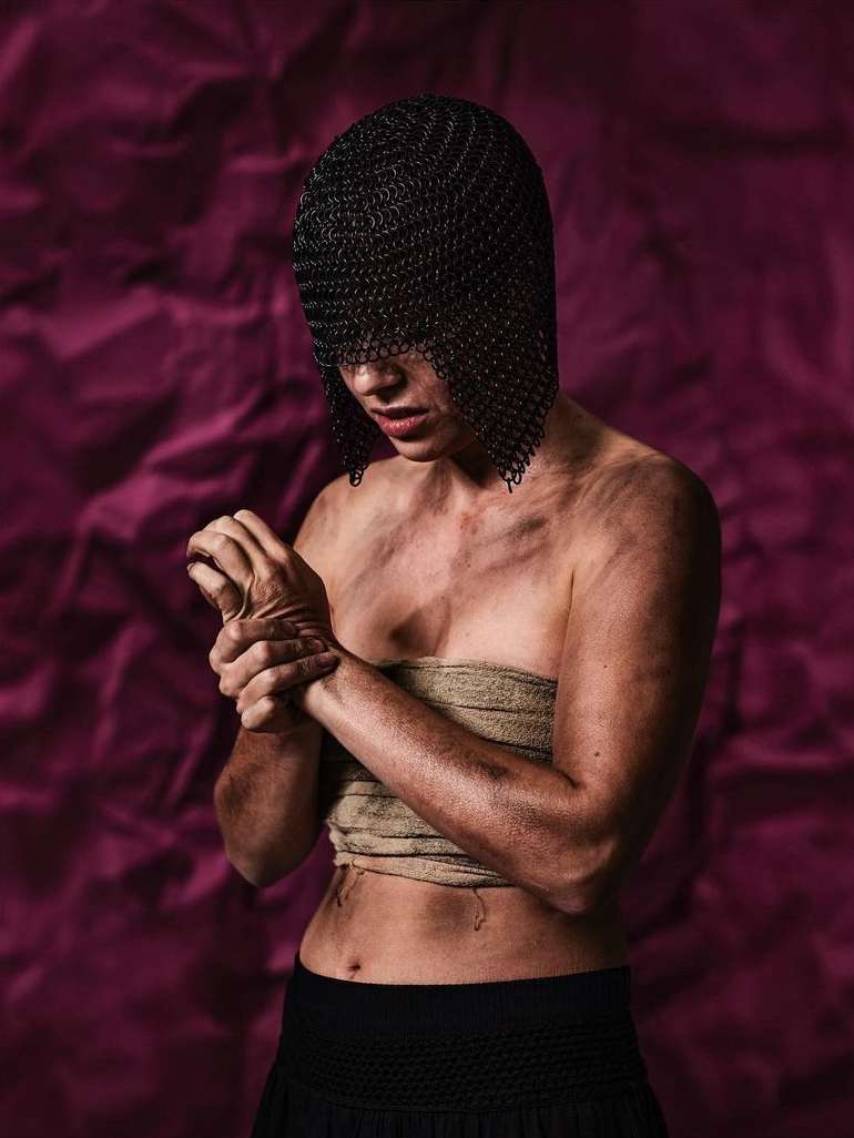 Woman in chainmail head covering and cloth wrap torso, gazing down with hands clasped against a textured purple backdrop.