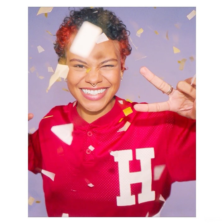 Close-up of a smiling person in a red jersey with short, curly hair, posing in front of a light purple background with confetti.