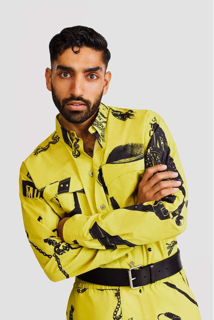 A man with a beard is standing in a yellow outfit with black prints on a light gray background. He has his arms crossed and is looking at the camera.