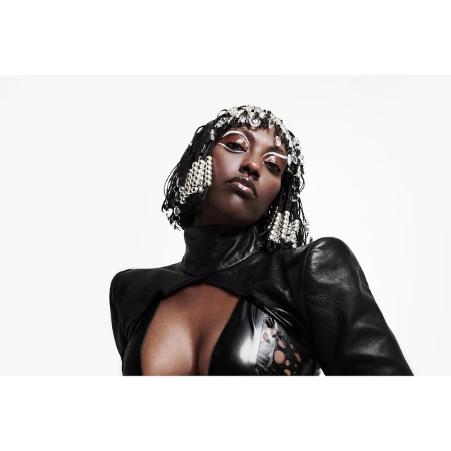 Striking close-up of a Black woman with decorative hairpiece and bold makeup, wearing a black top against a plain white background.