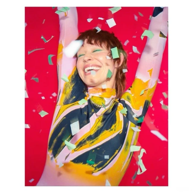 A woman with short brown hair and a patterned bodysuit smiles with confetti falling around her in front of a red background.