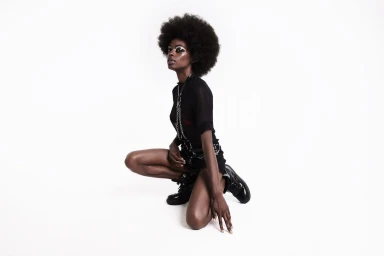 Black woman with an afro kneeling in black top and harness against a plain white background