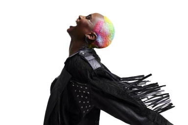 Black woman with rainbow dyed shaved hair and fringed black leather jacket laughing against a white background