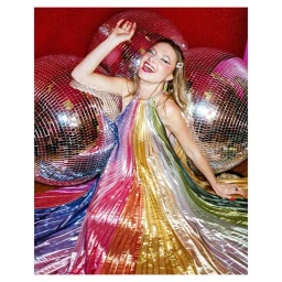 A smiling woman in a vibrant metallic rainbow pleated halter dress posing among large disco balls by Kate Bones.