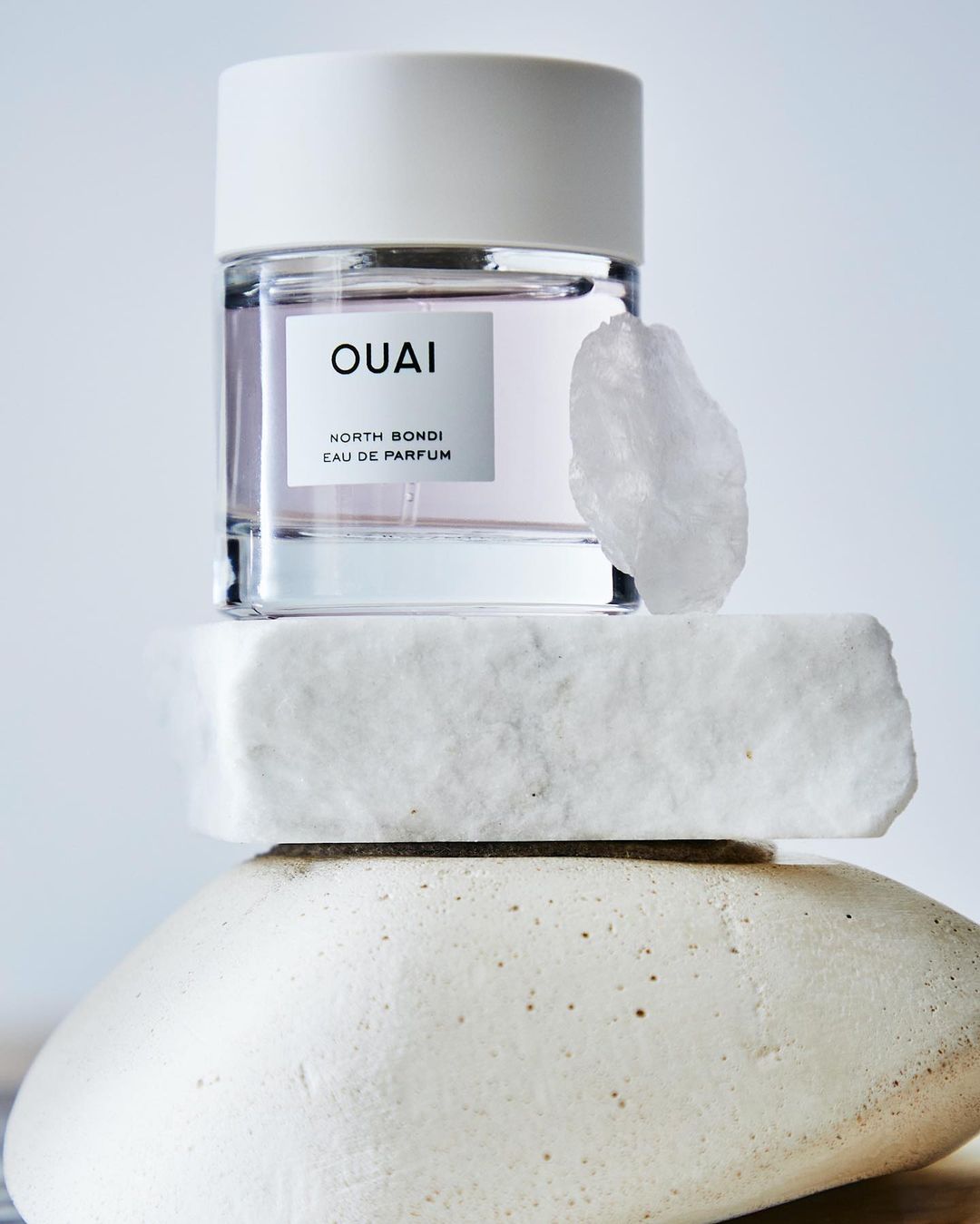 Product shot of OUAI North Bondi Eau de Parfum, a crystal, and layered stones against a light blue background in a vertical composition.