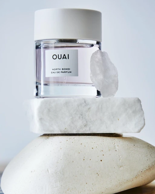 Product shot of OUAI North Bondi Eau de Parfum, a crystal, and layered stones against a light blue background in a vertical composition.