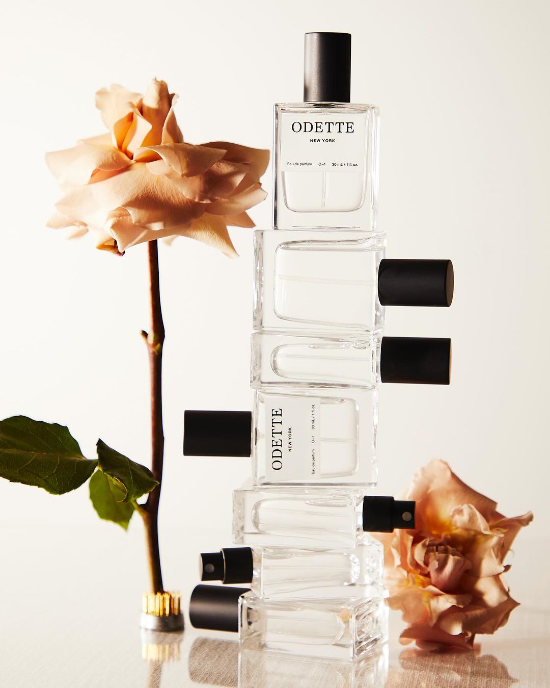 Stacked clear glass perfume bottles with black caps are displayed with beige roses, creating a still life product shot.