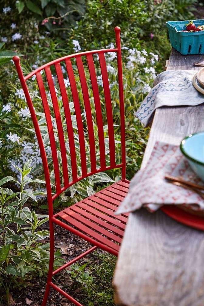 Red metal chair at a wooden table in a garden setting with flowers and greenery. Rustic outdoor dining scene, close up.
