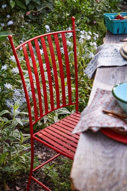 Red metal chair at a wooden table in a garden setting with flowers and greenery. Rustic outdoor dining scene, close up.