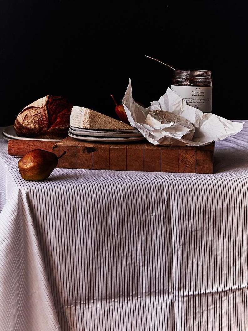 Still life photograph of a cheese selection, brown bread loaf, jam jar, and pears on a wooden cutting board and striped tablecloth.
