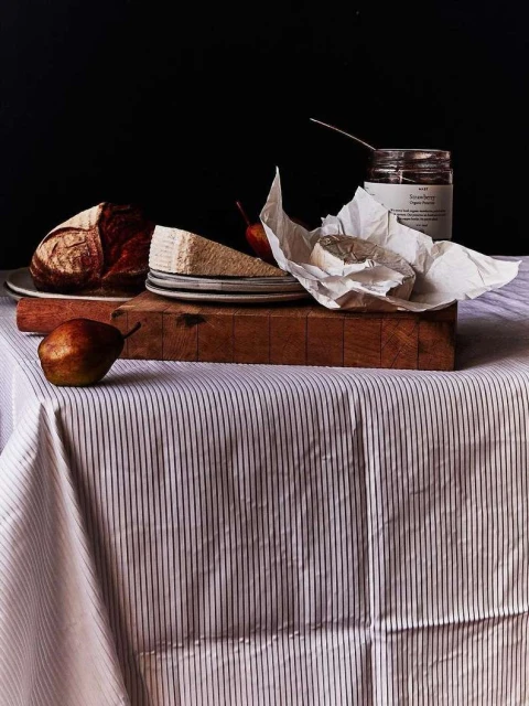 Still life photograph of a cheese selection, brown bread loaf, jam jar, and pears on a wooden cutting board and striped tablecloth.