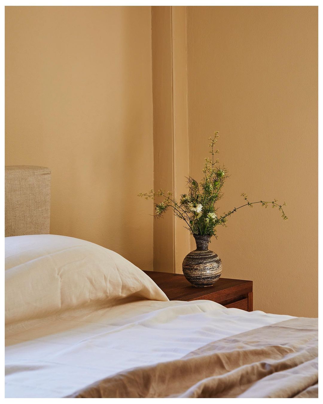 Bedroom interior features a bed with white and beige linens, and a dark wood nightstand holding a vase with white flowers.