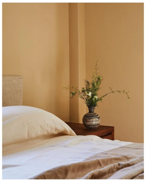 Bedroom interior features a bed with white and beige linens, and a dark wood nightstand holding a vase with white flowers.