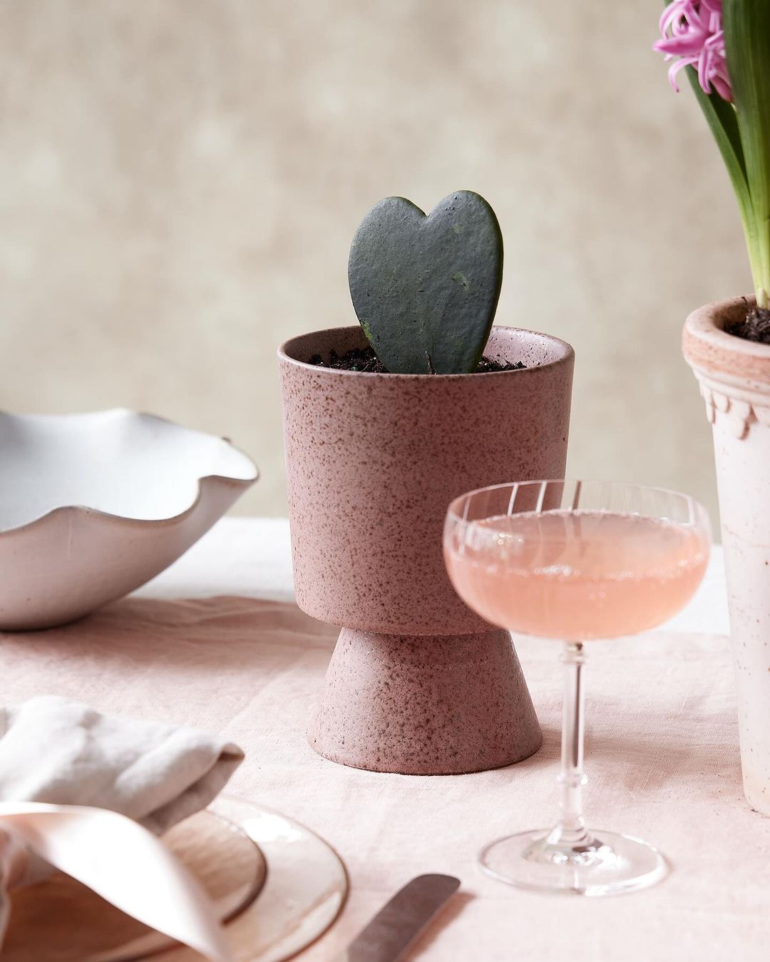 Close-up of heart-shaped hoya kerrii succulent in a pink pot on a table with a cocktail, white bowl, hyacinth plant and silverware.