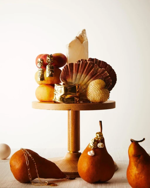 Still life composition featuring pears, peaches, a geode, and gold jewelry on a wooden cake stand, against a neutral backdrop.