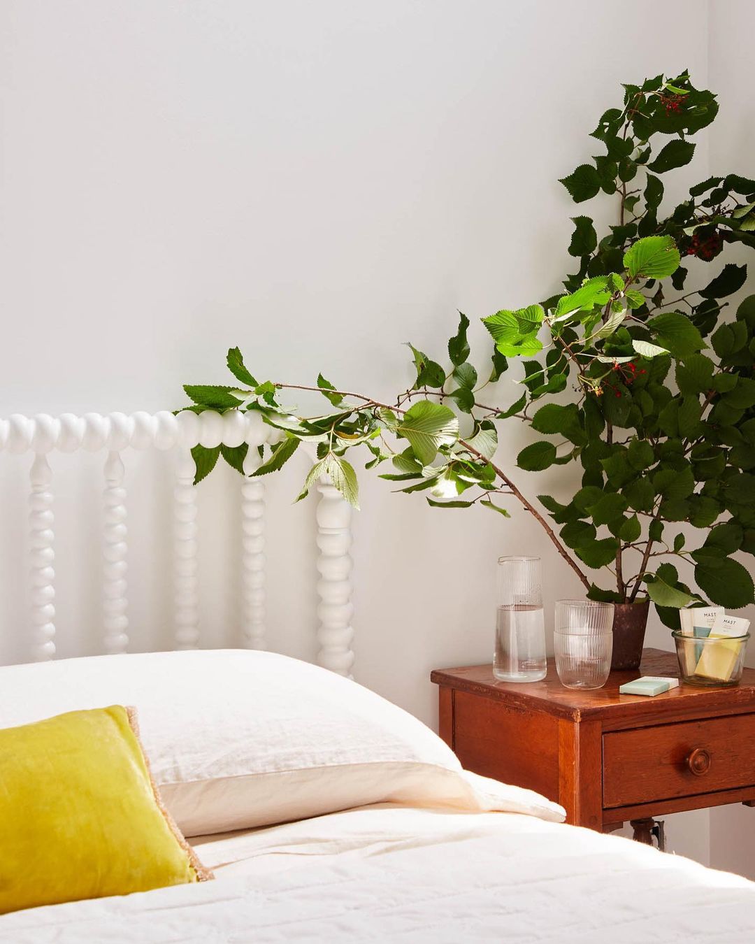 Bedroom interior with a bed with white linens and yellow pillow, a bedside table with a plant, water glasses, and bright natural light.
