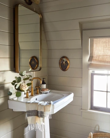 Bathroom interior with a white pedestal sink, gold faucet, a mirror, and linen window shade, creating a neutral, calming spa aesthetic.