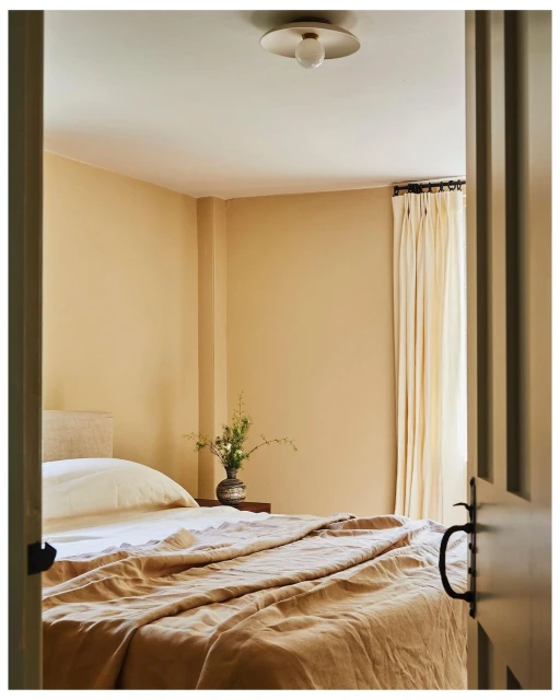 Bedroom interior featuring a neatly made bed with a light brown comforter, neutral beige walls, a wooden nightstand, and a modern ceiling light.