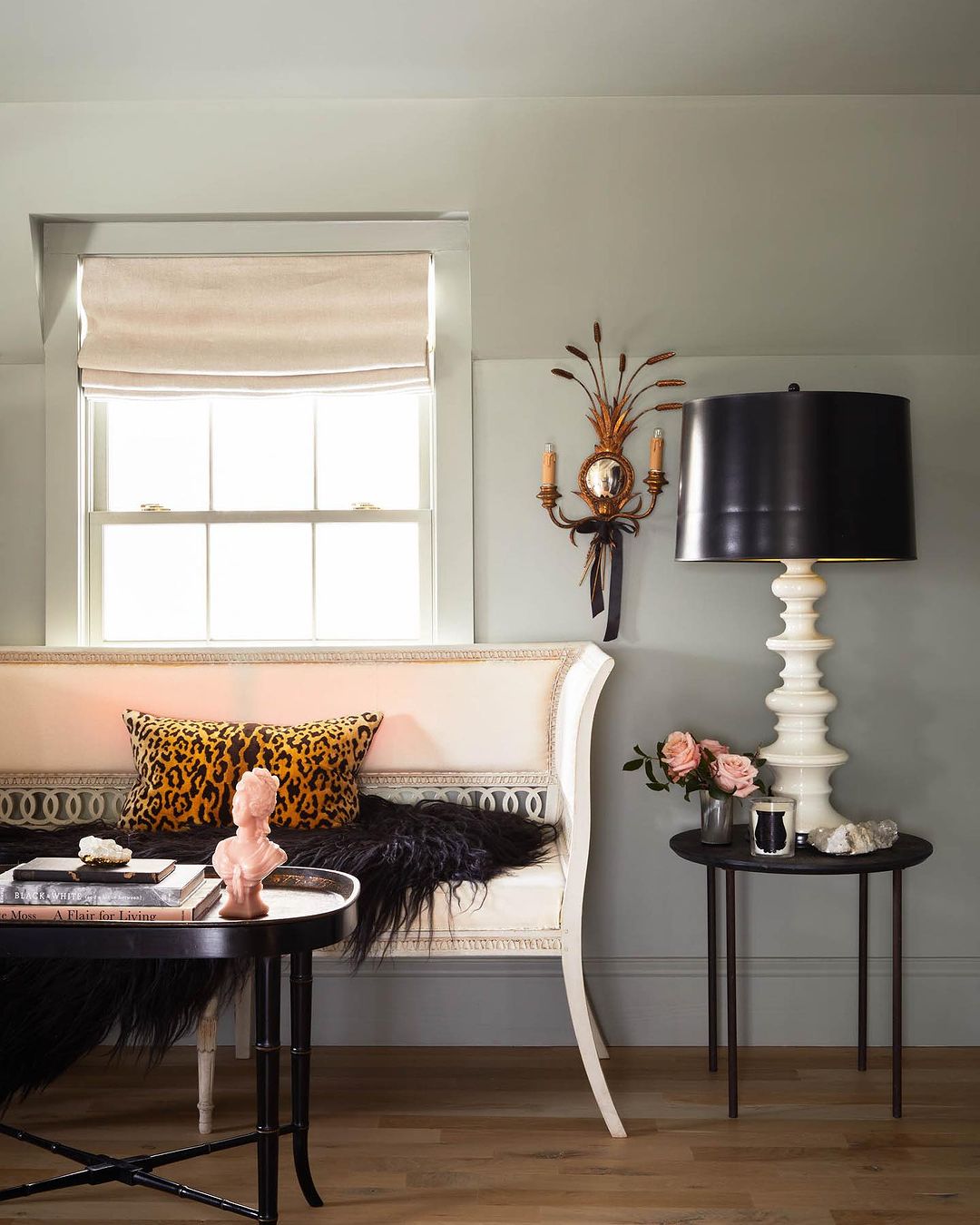 Interior view of living room with cream-colored settee, round tables, black lamp, flowers, and leopard print pillow in soft light.