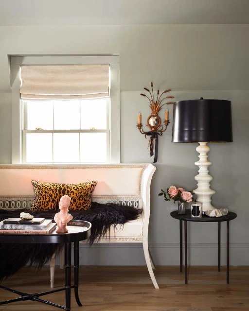 Interior view of living room with cream-colored settee, round tables, black lamp, flowers, and leopard print pillow in soft light.