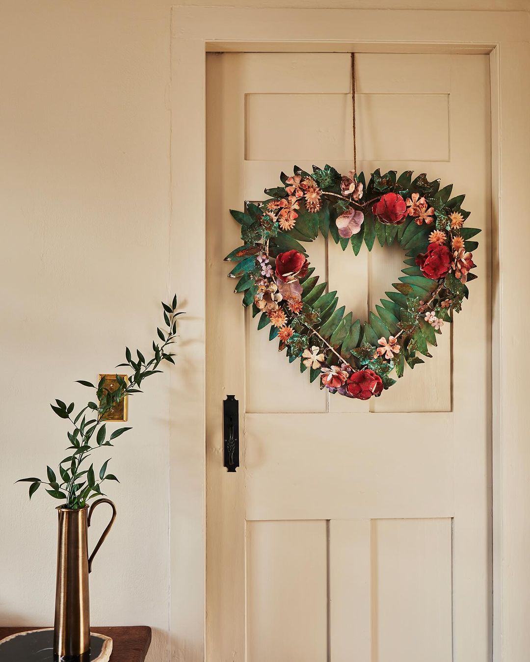 Heart-shaped floral wreath hanging on a white paneled door, featuring red and pink flowers, accompanied by greenery in a copper vase.