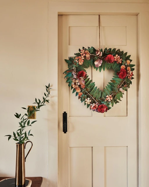 Heart-shaped floral wreath hanging on a white paneled door, featuring red and pink flowers, accompanied by greenery in a copper vase.