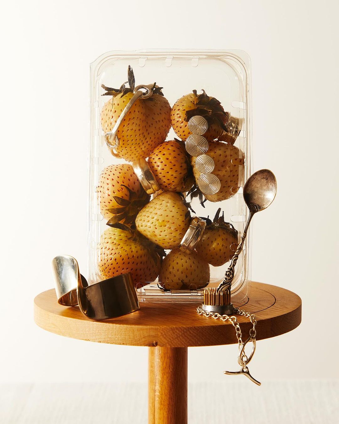 Strawberries in a clear plastic container on a wooden table, adorned with silver rings and a decorative spoon. White background.