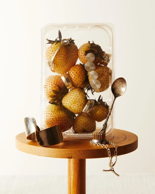 Strawberries in a clear plastic container on a wooden table, adorned with silver rings and a decorative spoon. White background.