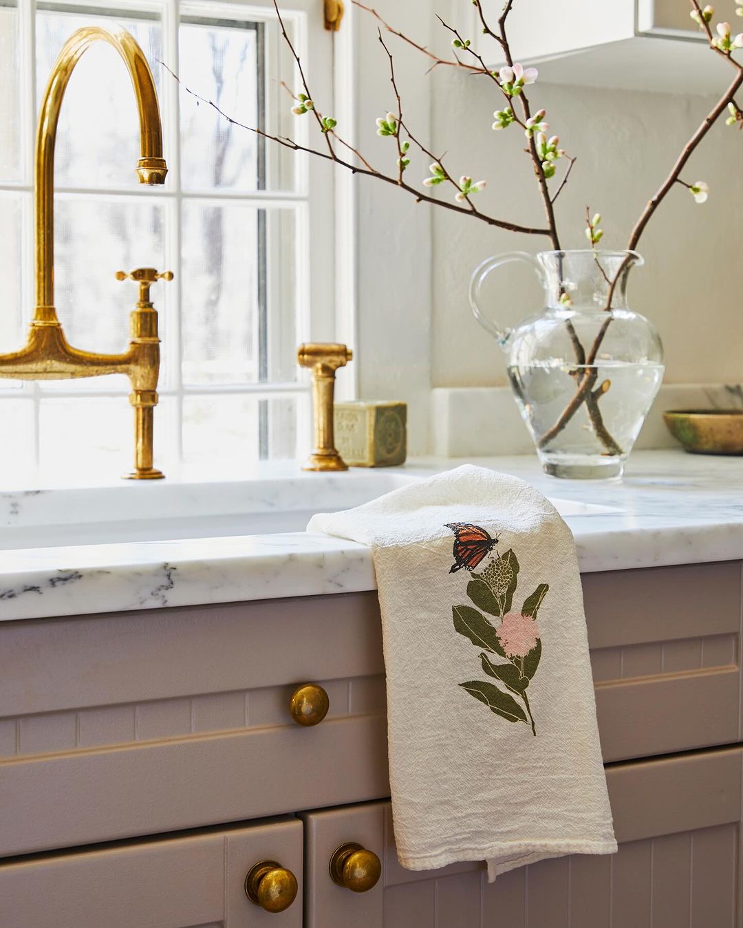 Interior view of a kitchen featuring a white embroidered butterfly tea towel draped over a gray cabinet, with a golden faucet and glass vase.