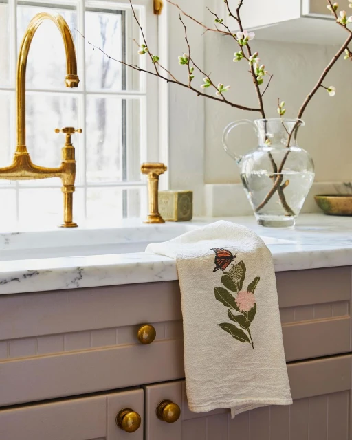 Interior view of a kitchen featuring a white embroidered butterfly tea towel draped over a gray cabinet, with a golden faucet and glass vase.