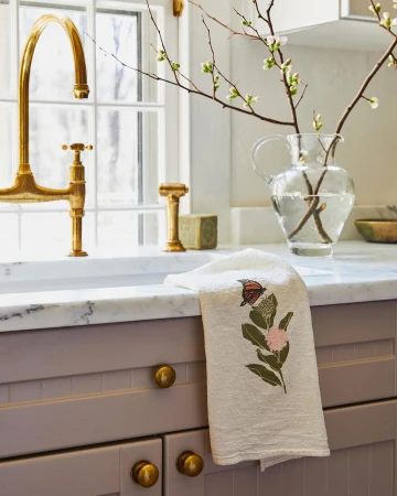 Interior view of a kitchen featuring a white embroidered butterfly tea towel draped over a gray cabinet, with a golden faucet and glass vase.