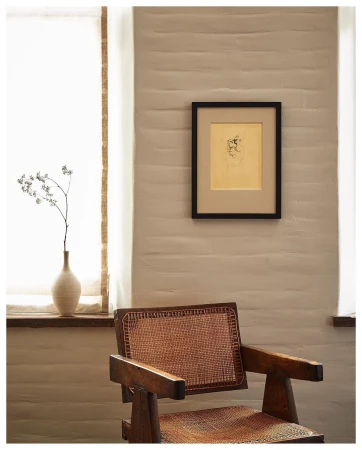 Framed sketch hanging on a white brick wall above a rustic wooden armchair with a woven cane seat.