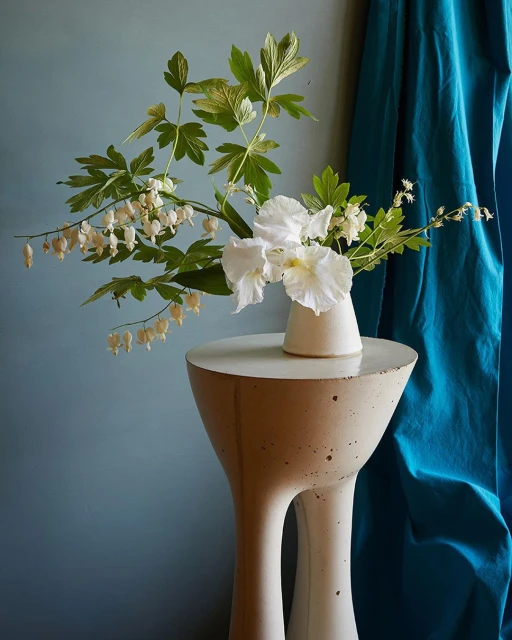 Floral arrangement featuring white flowers and foliage displayed on a contemporary sculptural table against a teal backdrop.