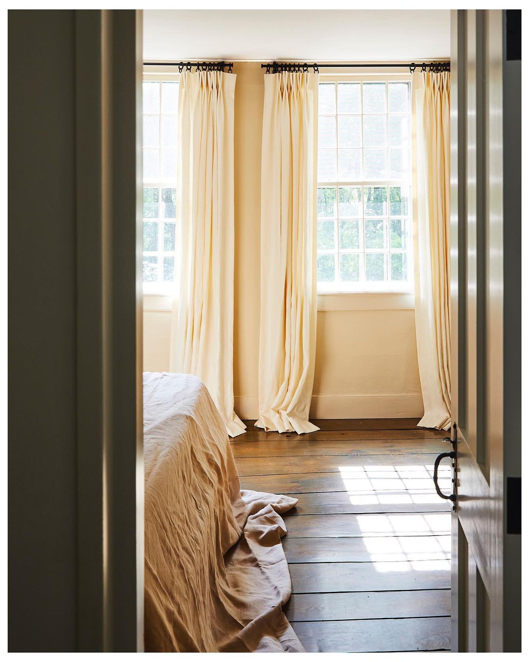 Bedroom interior featuring a neatly made bed with a peach-colored linen, windows dressed with cream-colored curtains, and hardwood floors.