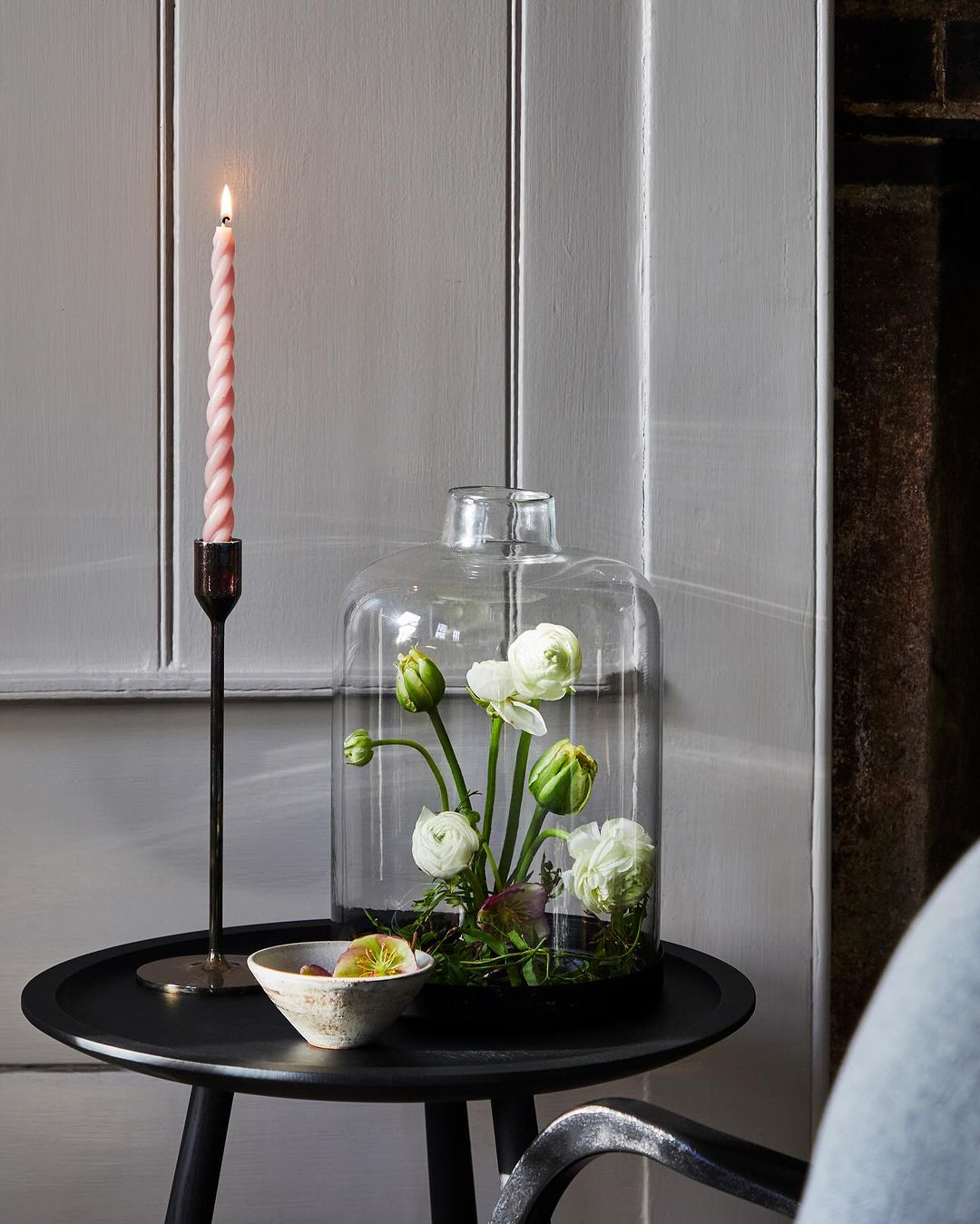 Still life shot of white ranunculus flowers in a glass cloche, a tall pink candle, and a small bowl on a round black table indoors.