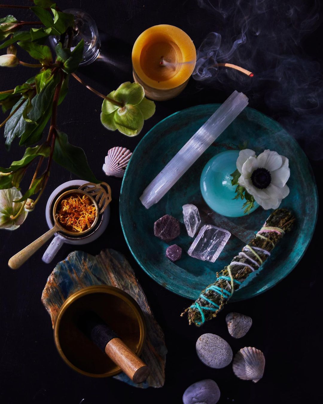 Still life of crystals, sage, candle, and flowers arranged on a plate on a dark surface, creating a spiritual and calming visual.