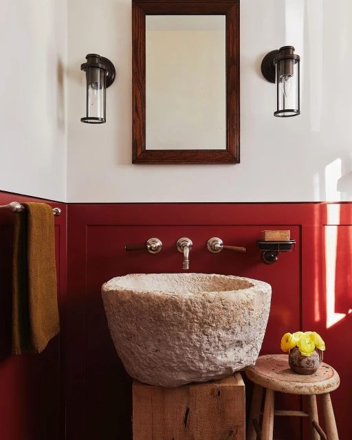 Rustic bathroom interior with a stone sink, dark red paneled wall, brown framed mirror, and sconce lighting creates a warm and inviting space.